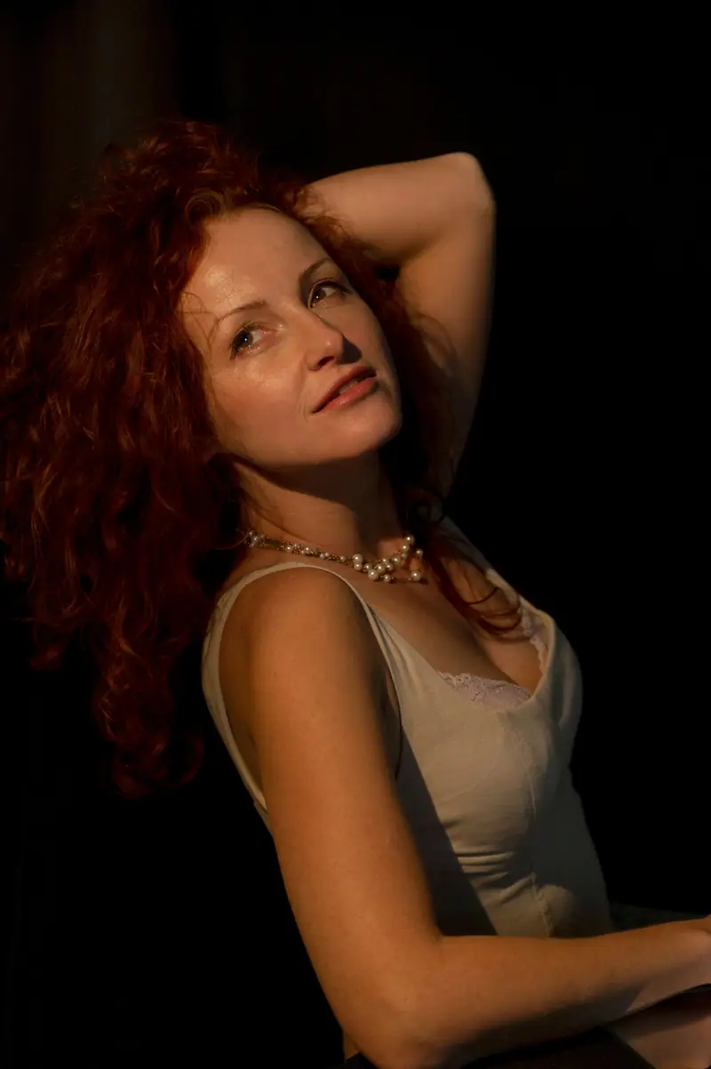 A woman with curly red hair in a light top against a dark backdrop